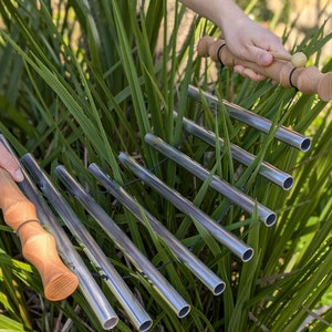 May include: A set of silver metal chimes with wooden mallets. The chimes are hanging from a metal frame and are surrounded by green grass.
