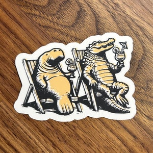 May include: A sticker featuring a cartoon manatee and alligator sitting in lounge chairs with tropical drinks. The manatee is light brown and the alligator is green. The background is a wood grain pattern.