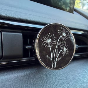 May include: A black car air freshener with a silver rim, featuring a white floral design of three daisies. The air freshener is round and sits on a car's dashboard, near an air vent.