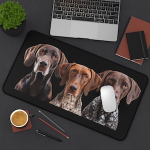 May include: A black and brown mouse mat featuring a design of three German Shorthaired Pointers. The dogs are looking at the camera. The mouse mat is on a black surface.