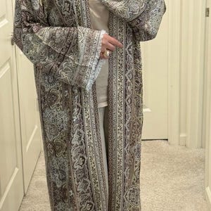 May include: A long, open-front duster cardigan featuring an intricate paisley pattern in shades of brown, beige, and cream. The garment has wide, long sleeves with frayed edges at the cuffs and hem, and a vertical border design along the front opening.