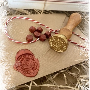 May include: A Christmas-themed craft kit featuring a wooden handled wax seal stamp with a gold-colored seal, red wax beads, and a red wax seal with a Santa Claus design. The kit includes a kraft paper envelope and red and white twine.