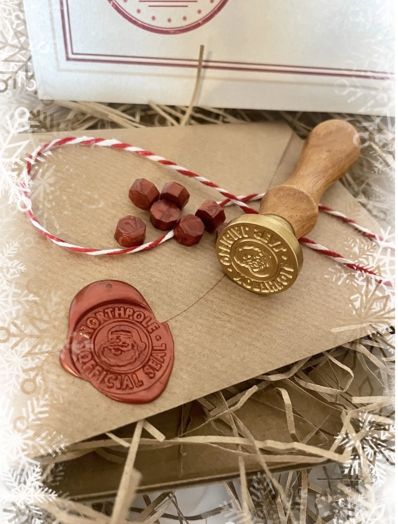 May include: A Christmas-themed craft kit featuring a wooden handled wax seal stamp with a gold-colored seal, red wax beads, and a red wax seal with a Santa Claus design. The kit includes a kraft paper envelope and red and white twine.