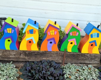 May include: Five colorful, whimsical birdhouses with unique, wavy designs. Each house is painted in a different bright color, including blue, yellow, orange, green, and yellow. The houses are arranged in a row on a wooden surface.