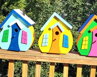 May include: Five colorful wooden birdhouses with unique designs. Each birdhouse features a different color scheme, including purple, blue, yellow, green, and pink. The birdhouses have a whimsical, cartoonish appearance with windows and a small entrance hole.