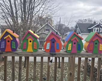 May include: A row of five colorful birdhouses with rounded bodies and pitched roofs. Each birdhouse features a unique combination of bright colors, including purple, green, orange, yellow, and pink. The birdhouses are mounted on a wooden railing.
