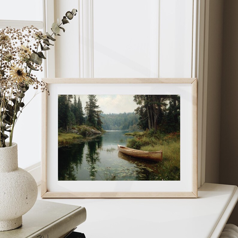 Peut inclure: Une impression encadr&eacute;e d'une sc&egrave;ne de lac paisible. L'&oelig;uvre repr&eacute;sente un bateau en bois flottant sur une eau calme, entour&eacute; d'une v&eacute;g&eacute;tation luxuriante et de grands arbres. L'&oelig;uvre est dans un cadre en bois clair, expos&eacute;e sur une surface blanche.