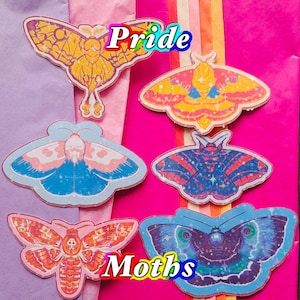 May include: A collection of colorful moth stickers with the words "Pride" and "Moths" in rainbow text. The stickers feature various designs, including yellow, blue, pink, and red moths. The background is a mix of pink, purple, and striped paper.