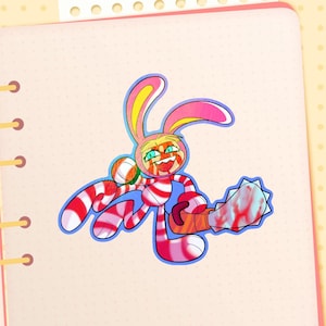 以下が含まれることがあります： ピンクと白のストライプのウサギがチェーンソーを持っている漫画のステッカー。ウサギは黄色の耳をしており、赤い飛沫で覆われています。チェーンソーは赤い飛沫のある青い刃を持っています。