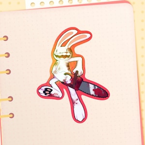 May include: A white cartoon rabbit sticker with a chainsaw. The rabbit has a menacing grin and is holding a bloody chainsaw. The sticker has a red outline and is on a pink background.