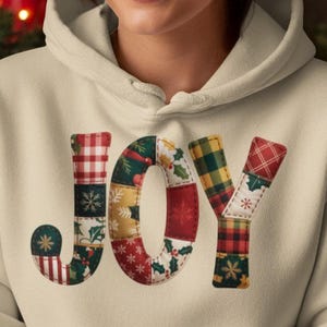 Könnte beinhalten: Ein beigefarbener Kapuzenpullover mit dem Wort "JOY" in Patchwork-Buchstaben. Die Buchstaben weisen verschiedene Muster zum Thema Feiertage auf, darunter Karos und Schneeflocken. Die Person hält eine rote Tasse.