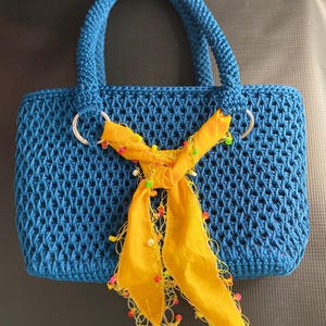 May include: A blue crocheted handbag with a rectangular shape and two handles. The bag features a decorative yellow scarf tied around the handles, adorned with colorful beads. The bag's dimensions are approximately 10 inches wide, 8 inches high, and 4 inches deep.