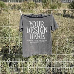 May include: A charcoal gray t-shirt hangs on a clothesline against a backdrop of a field and a cloudy sky. The shirt features the text "YOUR DESIGN HERE" in large white letters, with smaller text below that reads "THANK YOU FOR SUPPORTING REAL LIFE MOCKUPS."