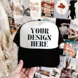 May include: A black and white trucker hat with a white mesh back. The front of the hat has the text "YOUR DESIGN HERE" in black.
