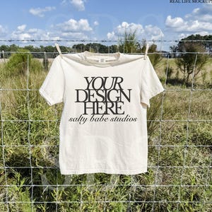 May include: A cream-colored t-shirt with the text "YOUR DESIGN HERE" and "salty babe studios" printed in black. The shirt is hanging on a clothesline against a backdrop of a wire fence, green grass, and a blue sky.