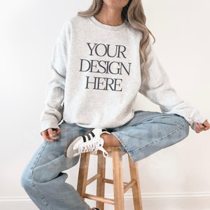 May include: A light gray sweatshirt with the text "YOUR DESIGN HERE" in black. The person is wearing light blue jeans and white sneakers with black stripes. The person is sitting on a wooden stool.