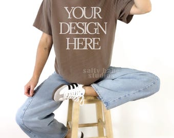 May include: A brown short-sleeved t-shirt featuring the white text "YOUR DESIGN HERE" printed on the chest. The shirt is paired with light blue denim jeans and white trainers with black stripes, worn while sitting on a wooden stool.