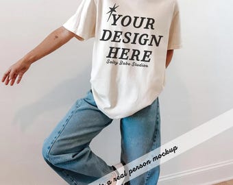 May include: An off-white short-sleeve t-shirt featuring the black text YOUR DESIGN HERE with a star icon above and the script text Salty Babe Studios below, paired with loose-fitting blue denim jeans and white sneakers with black stripes.