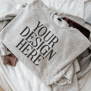 May include: A light gray sweatshirt with the text "YOUR DESIGN HERE" in large black letters. The sweatshirt is folded and layered with other sweatshirts in shades of gray and brown. A white hanger is visible.