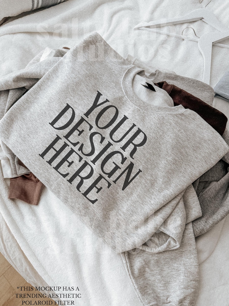 May include: A light gray sweatshirt with the text "YOUR DESIGN HERE" in large black letters. The sweatshirt is folded and layered with other sweatshirts in shades of gray and brown. A white hanger is visible.