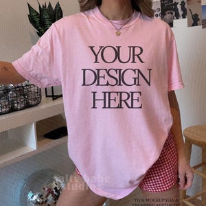 May include: A light pink t-shirt with the text "YOUR DESIGN HERE" in black. The shirt is paired with red and white checkered shorts. The image also includes text that says "THANK YOU FOR SUPPORTING REAL LIFE MOCKUPS" and "THIS MOCKUP HAS A TRENDING AESTHETIC POLAROID FILTER."