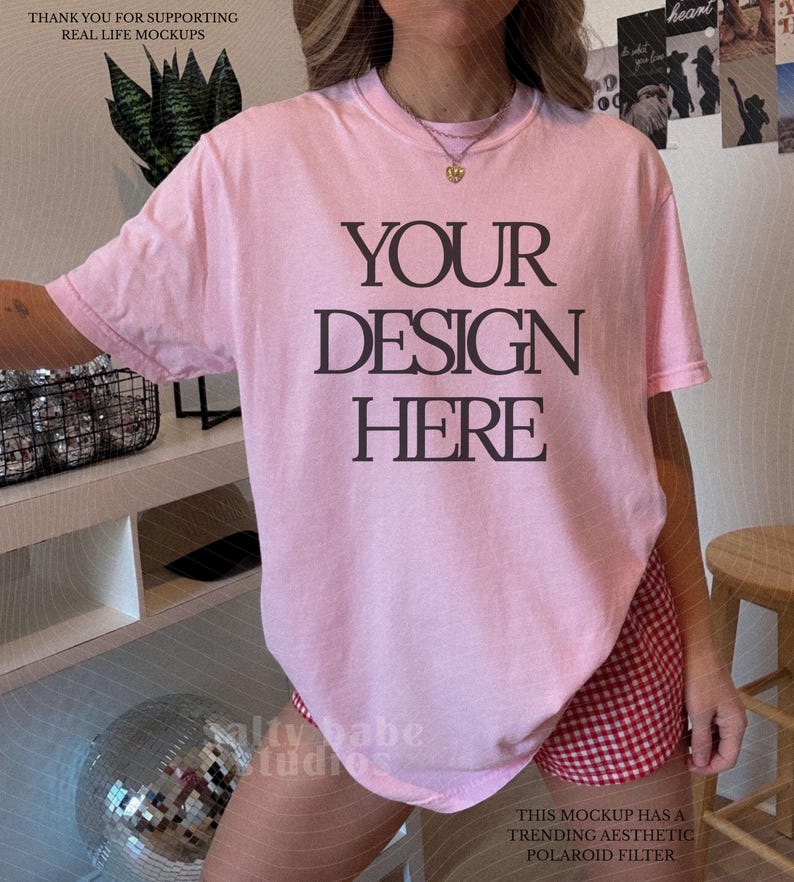 May include: A light pink t-shirt with the text "YOUR DESIGN HERE" in black. The shirt is paired with red and white checkered shorts. The image also includes text that says "THANK YOU FOR SUPPORTING REAL LIFE MOCKUPS" and "THIS MOCKUP HAS A TRENDING AESTHETIC POLAROID FILTER."