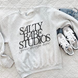 Op de afbeelding: Een lichtgrijs sweatshirt met de zwarte tekst "SALTY BABE STUDIOS your design here". Het sweatshirt wordt getoond met een blauwe jeans en witte sneakers met zwarte strepen.