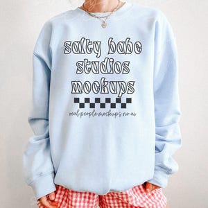 May include: A light blue sweatshirt with the text "salty babe studios mockups" in black lettering. The text is in a retro font style. Below the text is a black and white checkered pattern. The text "real people mockups no ai" is below the checkered pattern.