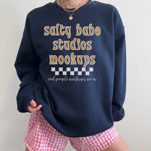 May include: A navy blue sweatshirt with the text "salty babe studios mockups" in gold lettering. The text "real people mockups no ai" is printed in white below the main text.