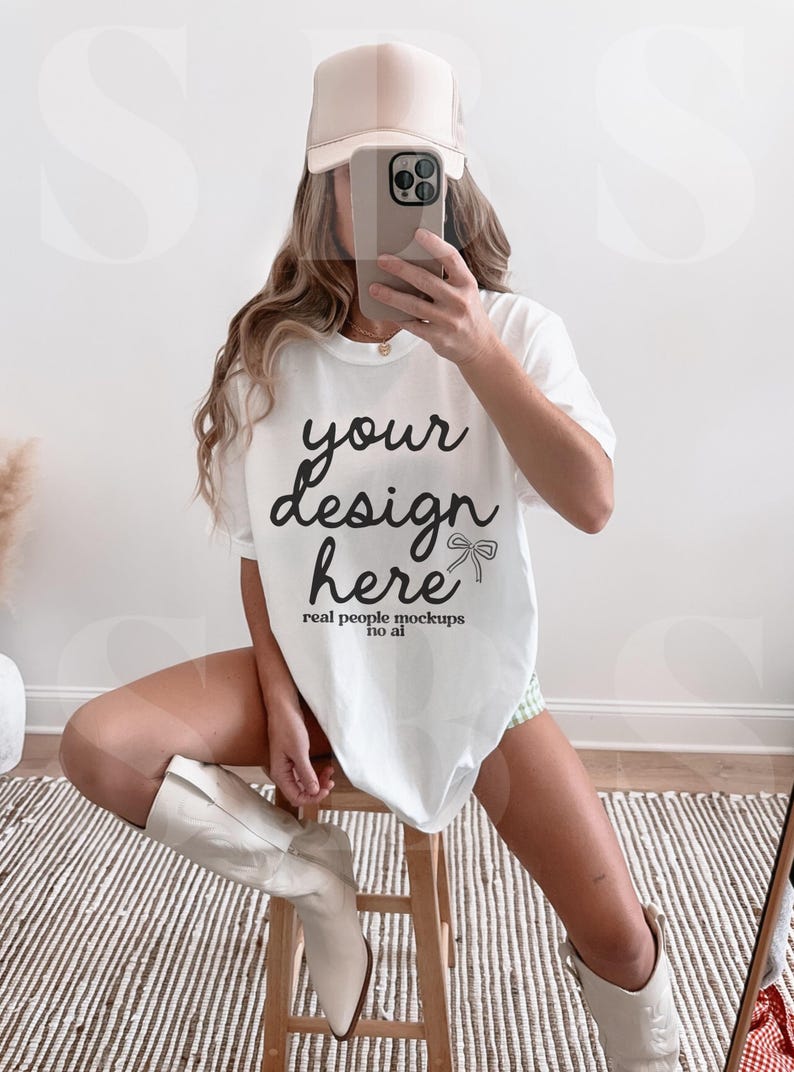 May include: A white oversized t-shirt with the text "your design here" and the words "real people mockups no ai" printed on the front. The model is wearing a beige baseball cap and white cowboy boots.