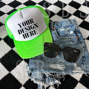 May include: A bright green and white trucker hat with a black mesh back. The hat has the text "YOUR DESIGN HERE" printed on the front. The hat is lying on a black and white checkered surface next to a pair of black sunglasses and a pair of blue denim shorts.