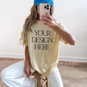 May include: A light yellow t-shirt with the text "YOUR DESIGN HERE" in black, paired with white pants and a blue cap. The image also includes a wooden stool and a smartphone.