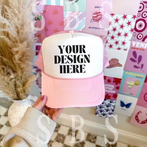 May include: Pink and white trucker hat with a white front panel and black text that reads "YOUR DESIGN HERE".