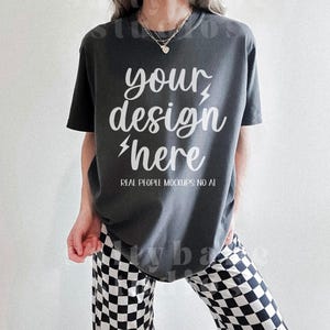 以下が含まれることがあります： 短い袖のチャコールグレーのオーバーサイズTシャツに、白い文字で「your design here」と書いてあります。シャツは、白黒のチェック柄のパンツを履いた人が着ています。デザインの下には、白い文字で「REAL PEOPLE MOCKUPS NO AI」と書いてあります。
