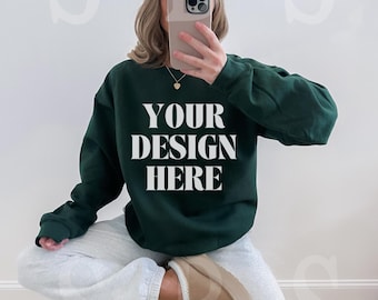 May include: A dark green crewneck sweatshirt featuring the white text YOUR DESIGN HERE printed on the chest. The garment is paired with light gray sweatpants, a beige baseball cap, and beige slip-on shoes, creating a casual ensemble.