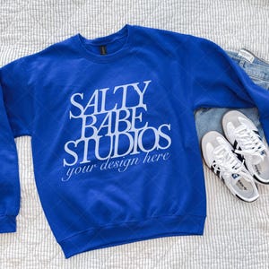 May include: Royal blue crewneck sweatshirt with the text "SALTY BABE STUDIOS your design here" in white. The sweatshirt is paired with light wash denim jeans and white sneakers with black stripes.