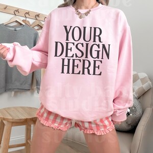 May include: A light pink sweatshirt with the text "YOUR DESIGN HERE" in black. The model is wearing pink and white plaid shorts with a ruffle trim. A wooden stool and a gray sweatshirt on a hanger are in the background.