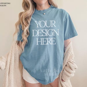Pode incluir: Uma camiseta azul com o texto "YOUR DESIGN HERE" em branco. A modelo está vestindo um cardigan bege e shorts brancos. A imagem é uma maquete para design de vestuário.
