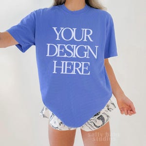 Può includere: Una maglietta blu con la scritta bianca "YOUR DESIGN HERE". La modella indossa pantaloncini fantasia. L'immagine è un mockup per il design di abbigliamento.