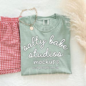 May include: A flat lay featuring a sage green t-shirt with the text "salty babe studios mockups real people no ai", red and white checkered shorts, and a gold chain necklace with a round pendant. A fluffy beige decorative plant is also present.