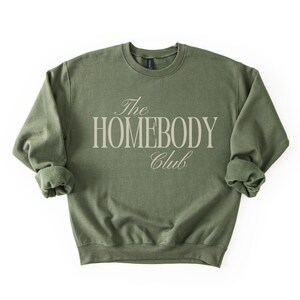 May include: An olive green sweatshirt with the text "The Homebody Club" in a light beige, elegant font. The sweatshirt has a crew neck and long sleeves with rolled cuffs. The garment is made of a soft material.