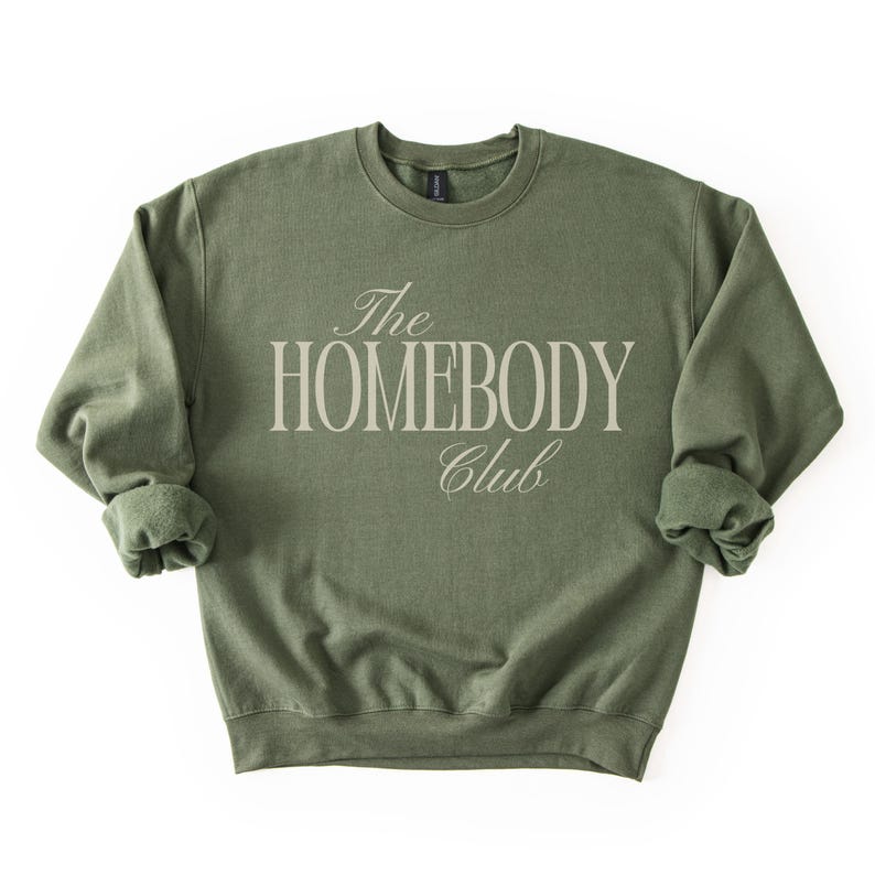 May include: An olive green sweatshirt with the text "The Homebody Club" in a light beige, elegant font. The sweatshirt has a crew neck and long sleeves with rolled cuffs. The garment is made of a soft material.