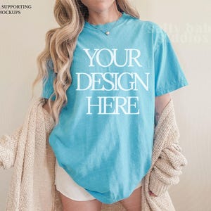 Puede incluir: Una camiseta turquesa con el texto "YOUR DESIGN HERE" en letras blancas. Un cárdigan beige está drapeado sobre los hombros. La imagen es una maqueta para el diseño de ropa.