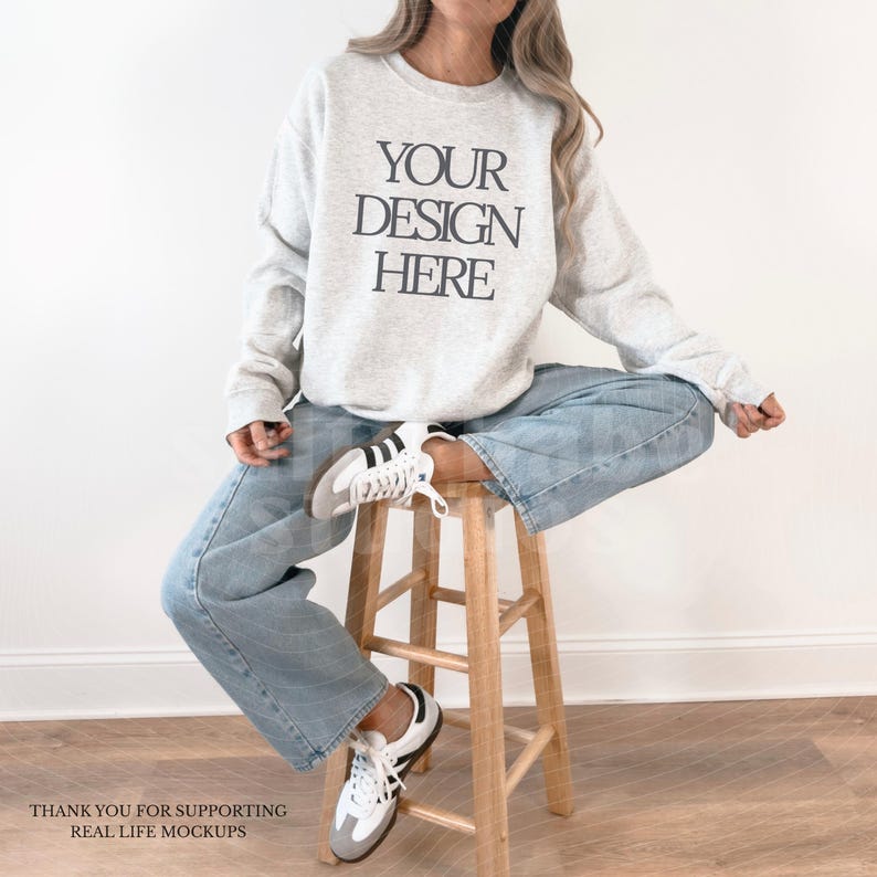 May include: A light gray sweatshirt with the text "YOUR DESIGN HERE" in black. The person is wearing light blue jeans and white sneakers with black stripes. The person is sitting on a wooden stool.