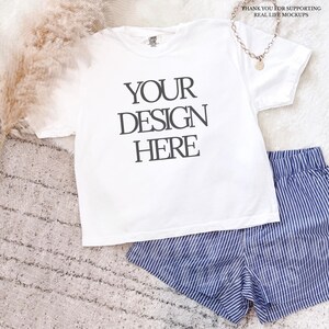 Pode incluir: Uma camiseta branca com o texto "YOUR DESIGN HERE" em preto, combinada com calções listrados em azul e branco. Um colar dourado com um pingente e o texto "THANK YOU FOR SUPPORTING REAL LIFE MOCKUPS" também são visíveis.