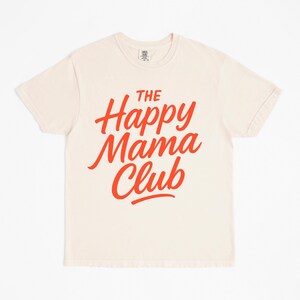 May include: Cream-colored t-shirt with the text "The Happy Mama Club" in bright red, stylized lettering. The shirt has short sleeves and a crew neck. The fabric appears to be soft and comfortable.