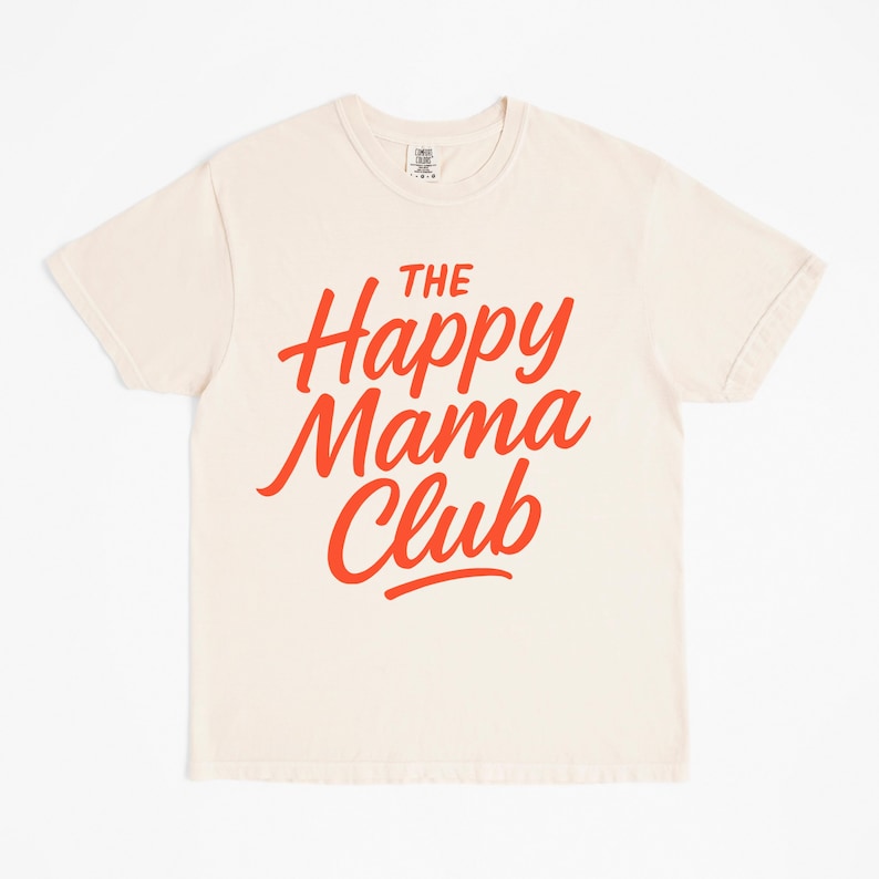 May include: Cream-colored t-shirt with the text "The Happy Mama Club" in bright red, stylized lettering. The shirt has short sleeves and a crew neck. The fabric appears to be soft and comfortable.