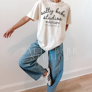 May include: Cream-colored t-shirt with the text "salty babe studios mockups real people no ai" in black script. The shirt is paired with light blue denim jeans and white sneakers with black stripes.