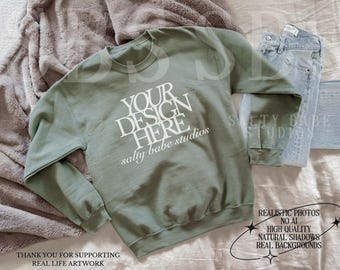 May include: A sage green crewneck sweatshirt featuring the white text YOUR DESIGN HERE and salty babe studios printed on the front. The garment has long sleeves, a ribbed collar, and ribbed cuffs, presented as a blank canvas for custom graphics.