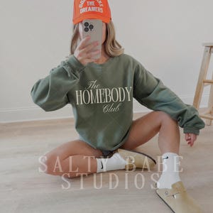May include: An olive green sweatshirt with "The Homebody Club" printed on the front. The image also shows an orange cap with the text "Don't Wake The Dreamers". The person is wearing tan clogs and white socks.
